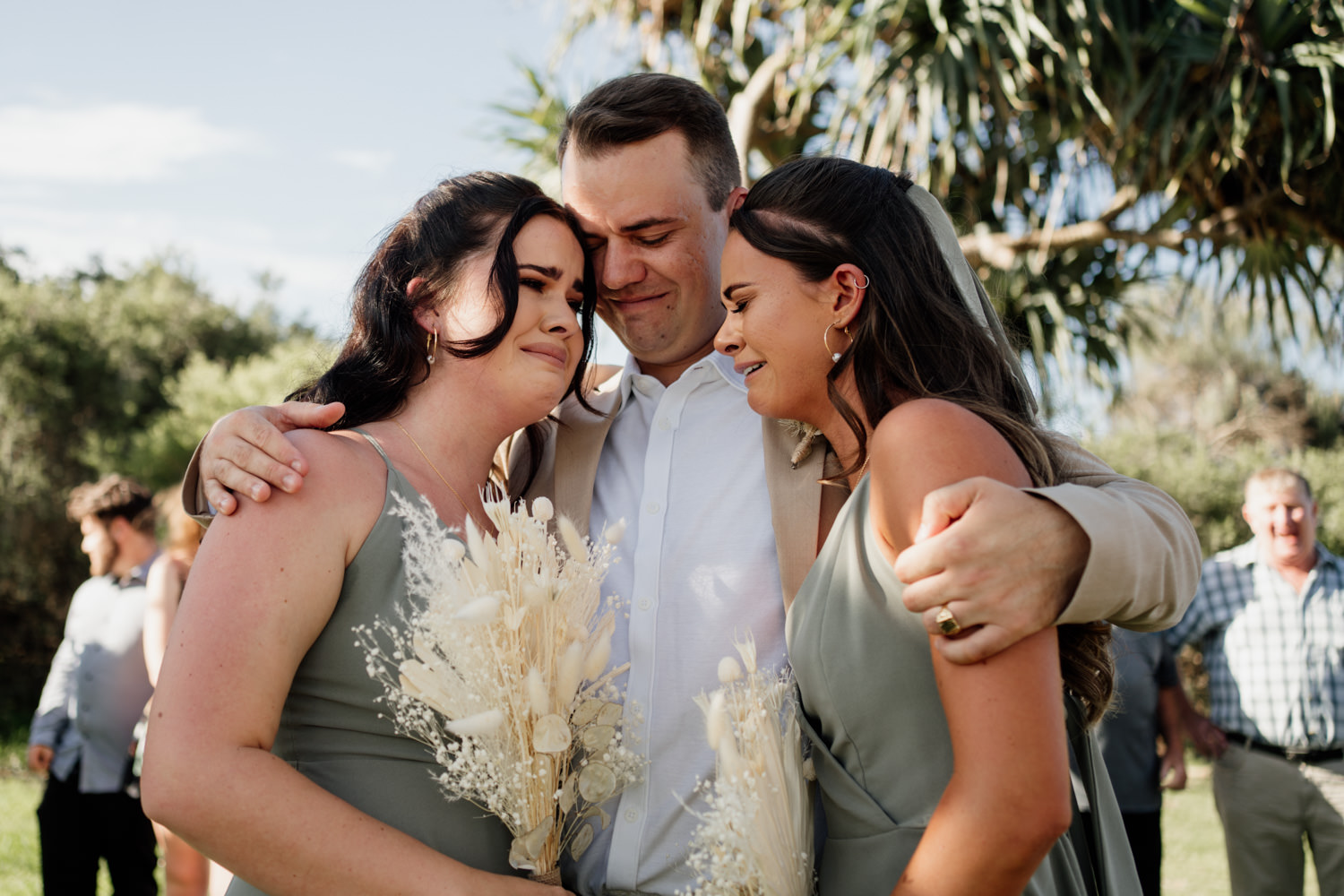 Groom and his two sisters hugging and crying post ceremony by Byron Bay wedding photographer Sam Wyper